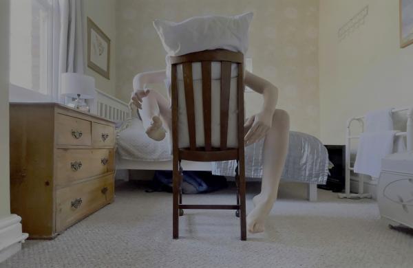 A figure in a bedroom sits backwards on a chair with a pillow covering their head and limbs contorted.