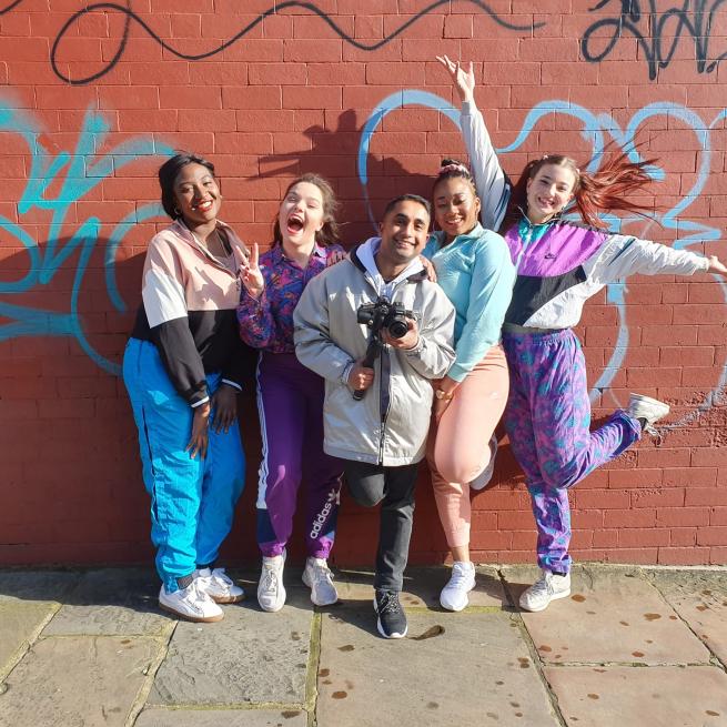 Four female dancers and a photographer posing in front of a red brick wall