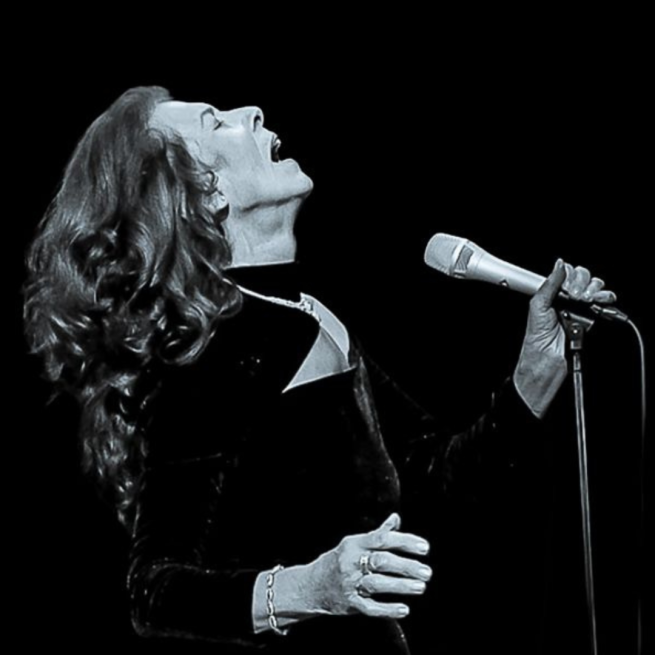 Black and white image of Elkie Brooks singing into a microphone on a stand with a plain black background.