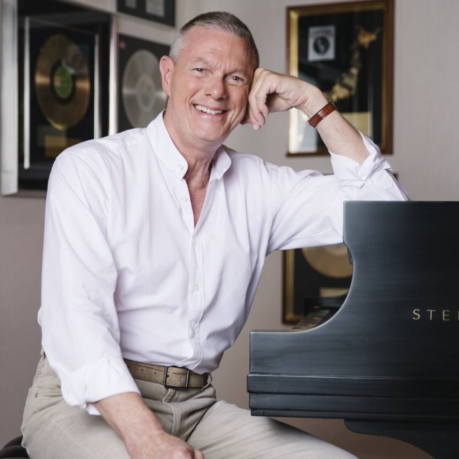 Richard Carpenter wearing a white shirt smiles to the camera whilst leaning on a piano.