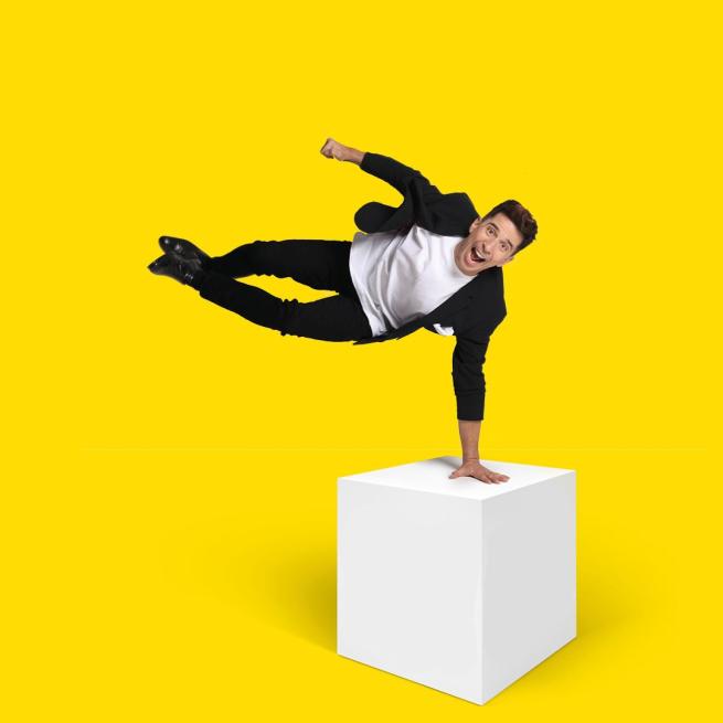 Russell Kane balancing on one arm on a white box with his legs in the air on a yellow background.