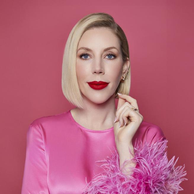 Katherine Ryan with deep red lipstick on and a pink silk top with a fluffy sleeve. Her left hand is brought up near her face and she is against a coral background.