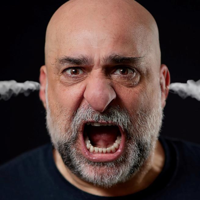 Omid Djalili yelling at the camera with steam coming out of his ears. All on a black background