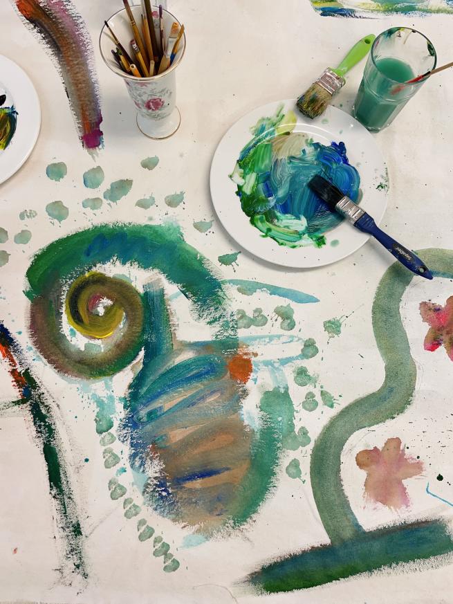 Painting of swirls alongside a pot of pencils, a plate covered in paint and brushes