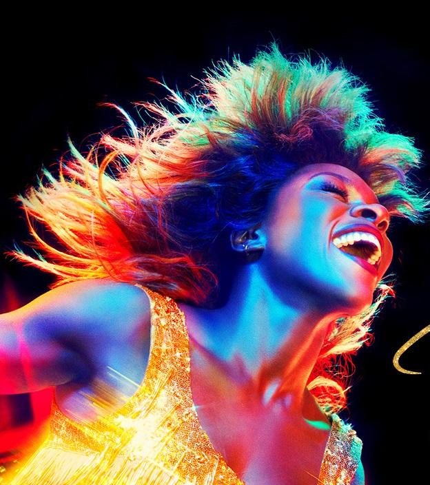 Image of Tina Turner with a rainbow tinge to the image. Tina is smiling wide in a gold fringe dress. 
