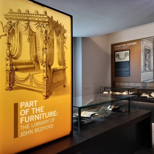 Yellow illuminated exhibition panel which reads "Part of the Furniture: The Library of John Bedford" and has an illustration of a four poster bed, behind which can be seen glass museum cases filled with books.