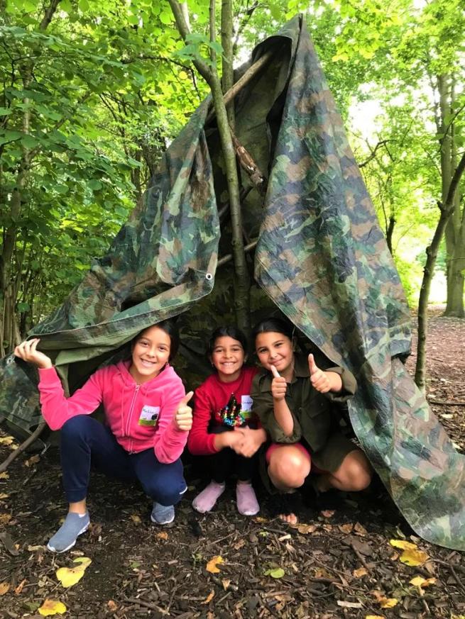 Photo shows three girls in a tarpaulin shelter that they built in the woods.