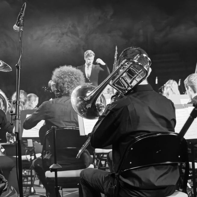 Black an white picture from the back of an orchestra with a trombonist as the main focus and the conductor in the background.