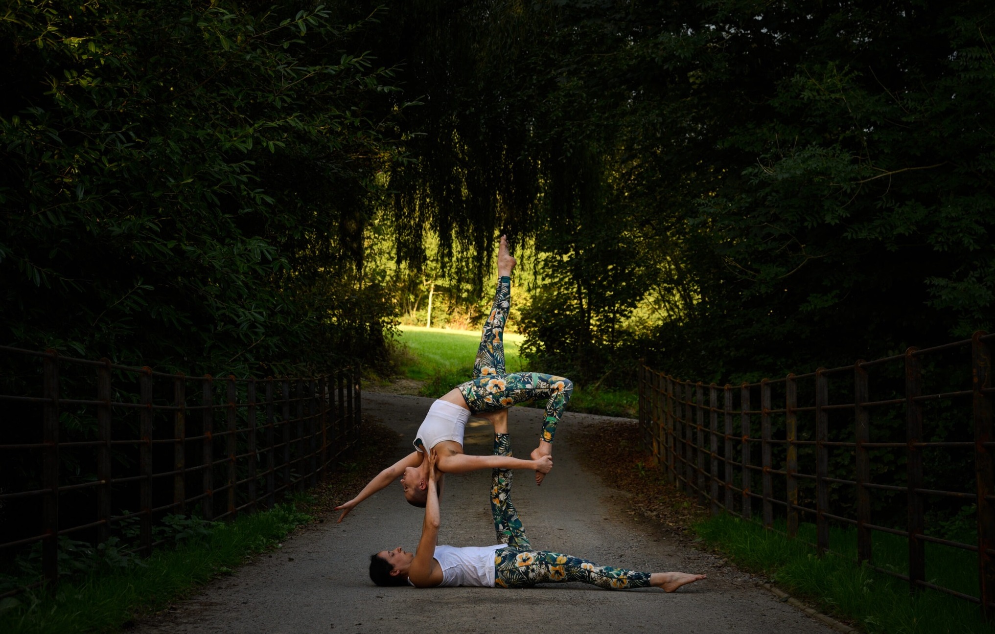 2 women in a park pathway, covered by trees. Theyr'e wearing yoga clothing and are balanced, one holding up the other in the air, who is arched back and left leg up in a striking pose