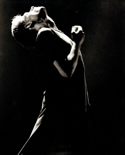 Black and white image of a young man mid 20's head thrown back looking at the sky face and chest lit by light from above his right arm bent with his hand in front if his chest.