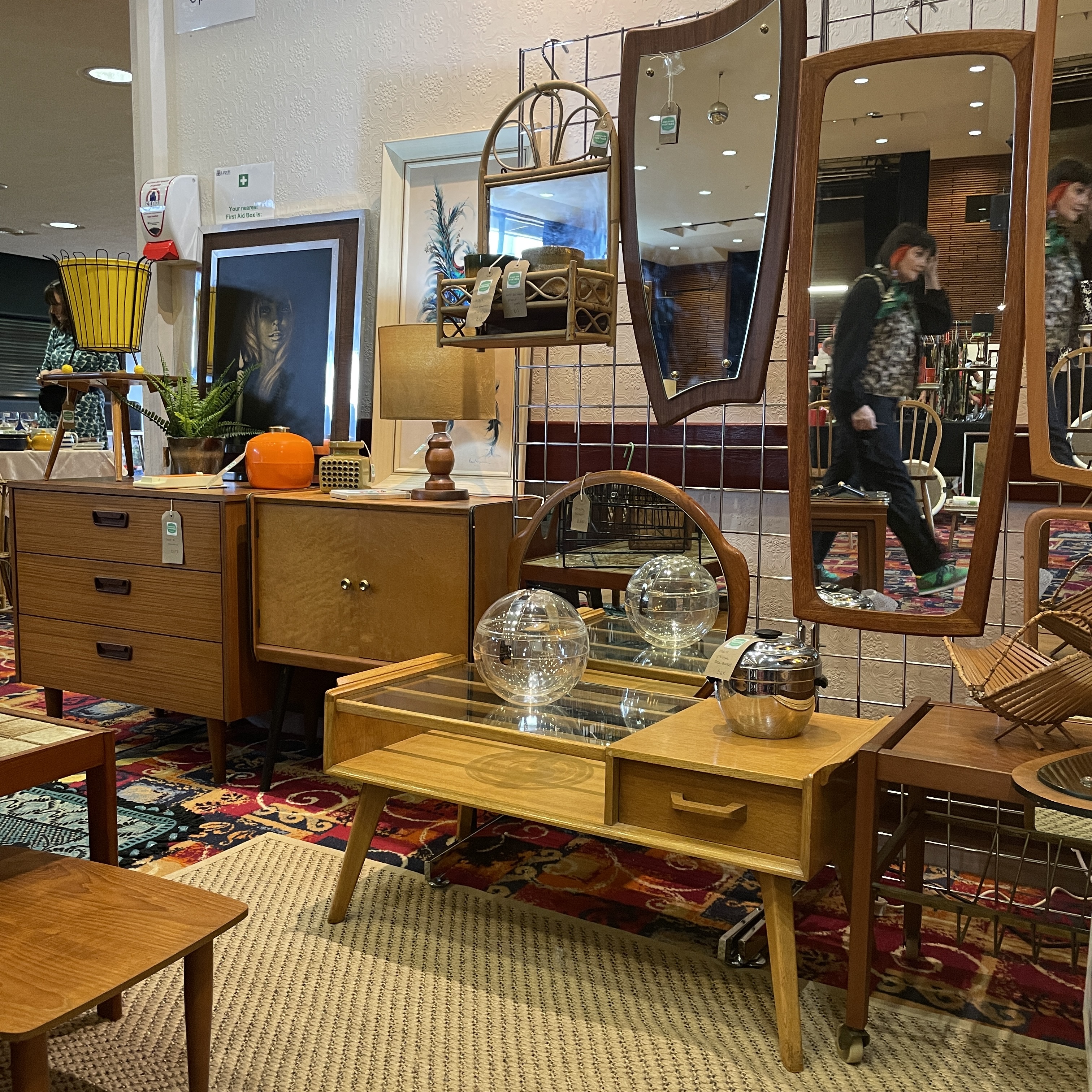 Teak framed mirrors on a wall. A display of vintage tables and cabinets. Lamps and vases on tables.