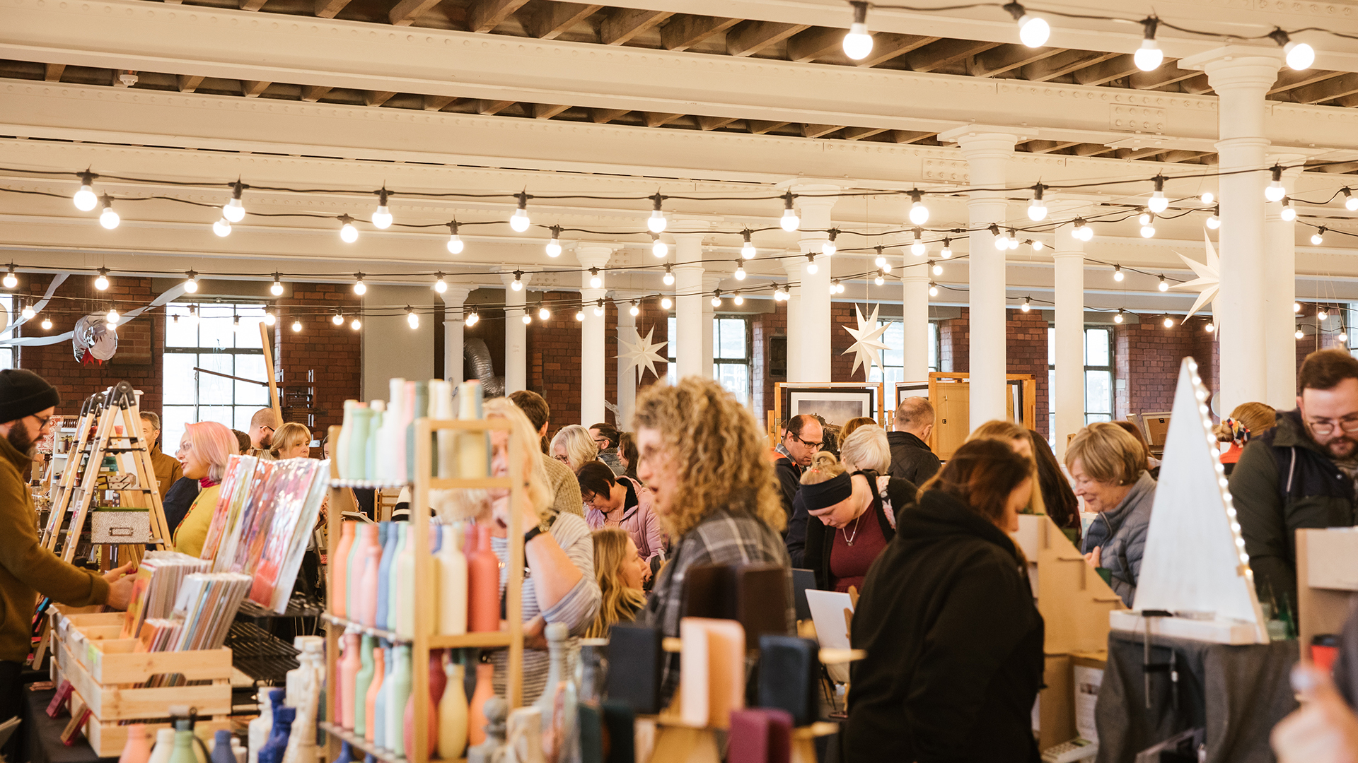 A previous Sunny Bank Mills Christmas Market in full swing in our 1912 Mill. The space is a former textile mill, with high beamed ceiling and tall white pillars in the centre. The space is filled with strings of lights and christmas decorations and visitors are shopping at a range of stalls stocked with handmade items including ceramics and prints.