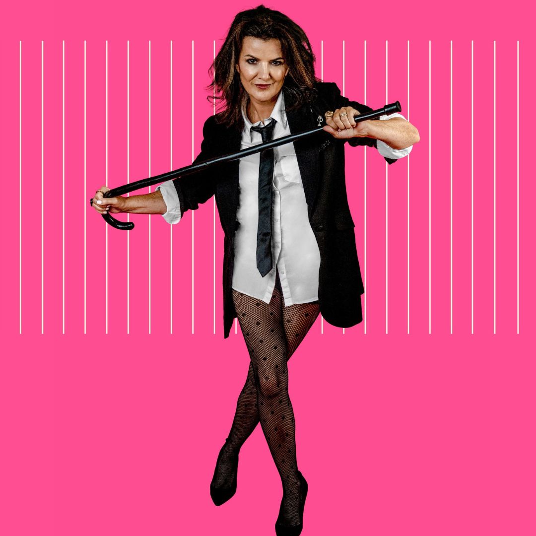 Deirdre O'Kane in a suit jacket, black tie, white shirt and just semi-sheer tights and black heels holding a cane across her chest with both hands stood against a pink background.