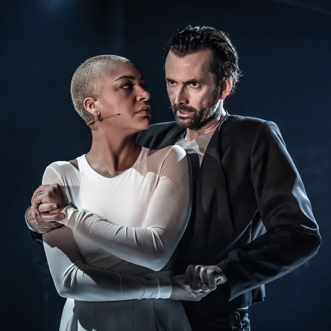 David Tennant as Macbeth in a suit jacket and grey t-shirt looking stern holding on to Cush Jumbo as Lady Macbeth wearing a white dress with shoulder cut outs who is looking back at David.