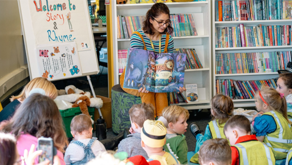 A colourfully dressed woman sits in front of a bookshelf reading a large picture book to a group of children.