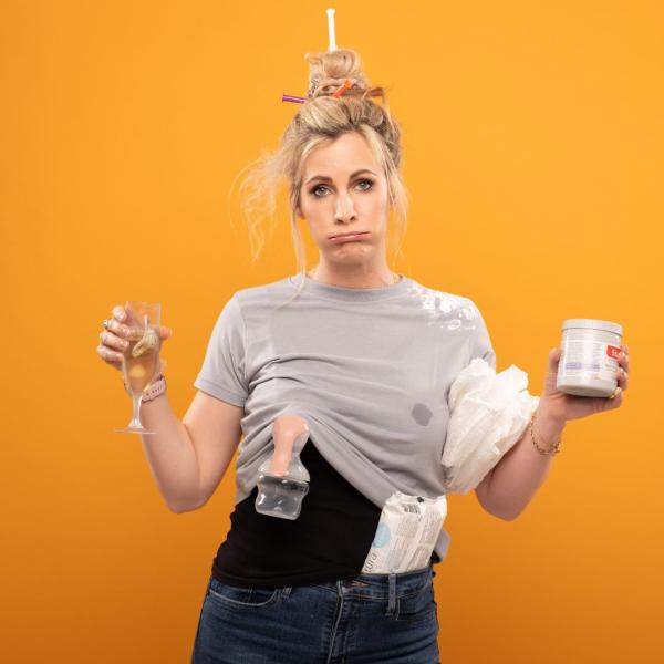 Hannah East stands sighing and exasperated with a messy bun, her arms outstretched holding a prosecco flute with a dummy floating in it, and a tub of Sudocrem. Her shirt is stained and a breast milk pump is on display.
