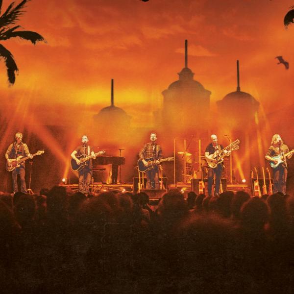 In front of a hazy, deep orange skyline of buildings, palm trees and a soaring eagle, six guitarists perform on stage to a crowd.
