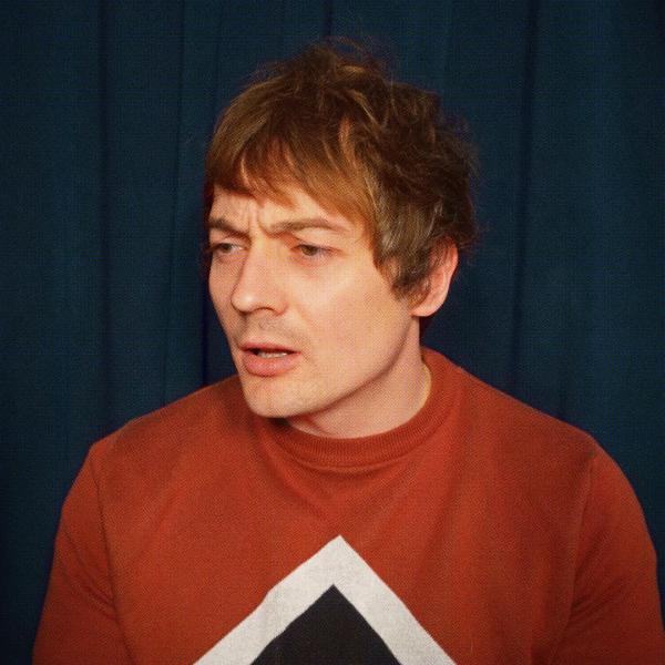 Matt Bragg squints with his mouth partially open, looking off camera in an orange jumper in front of a navy curtain backdrop.