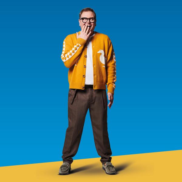 Alan Carr poses with his hand covering his mouth and cheeky expression, wearing a mustard-yellow cardigan. He stands in front of a bright blue backdrop.