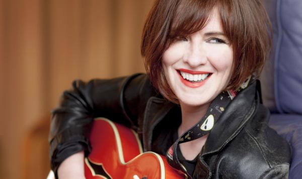 A lady sits with guitar she is wearing a black leather jacket and has bouncy auburn hair a wonderful smile and red lipstick