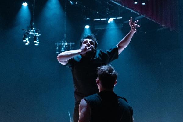 On a dimly lit stage, the camera is looking up at 2 male dancers dressed in black. One has his back to the camera and is holding up the other male performer. The one being lifted is facing the camera, with his right hand over his mouth as if stopping himself from speaking. His left arm is reaching out as if to try and grab something.