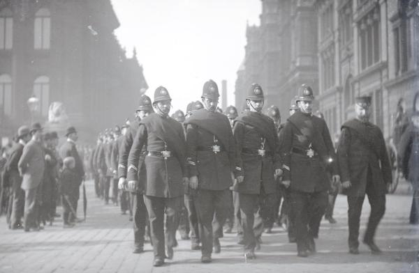 Black and white picture of Victorian policemen marching in formation while pedestrians look on. To the left is Leeds Town Hall. Picture taken in the Leeds Gas Strike of 1890.