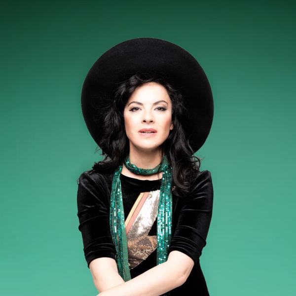 Camille O'Sullivan wearing a big black hat and green sparkly scarf sat in front of a green background.