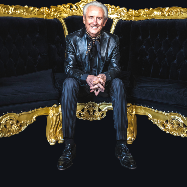 Tony Christie in a black leather suit jacket and black and gold neck tie sat on a black and gold sofa.
