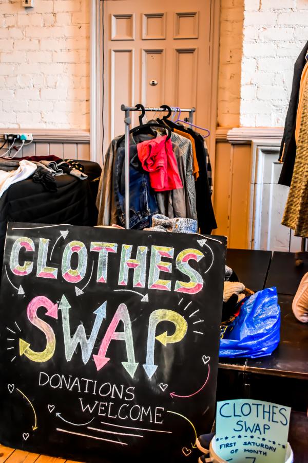 A chalkboard wih the words 'Clothes Swap, Donations welcome' written on in coloured chalk. Behind the sign are rails of clothes.