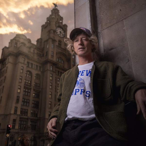 John Power looking down at the camera wearing a camo bomber jacket leaning against a concrete wall with a tall concrete building in the background.