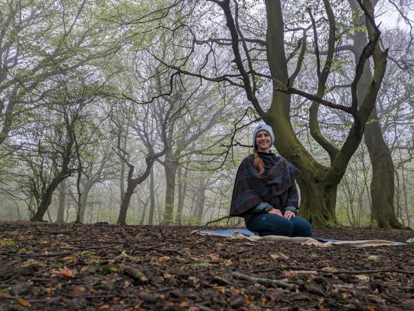 A photograph of Alice sat on a yoga mat within woodland. 