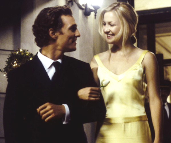 Man in suit and blonde woman in yellow evening dress link arms and look into each other's eyes, smiling.