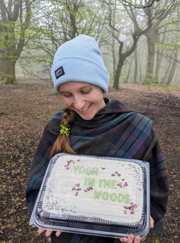 A photograph of Alice holding a Cake with Yoga in the Woods written on it, set within Woodland
