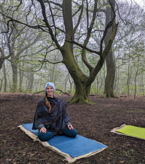 A photo of Alice sat in an open posture on a yoga mat set within Woodland