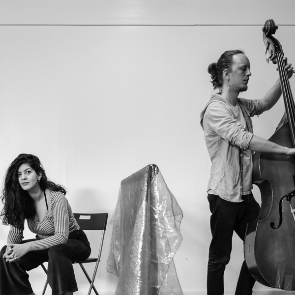 Black and white photograph of a woman sat on a folding chair next to a man stood playing a cello