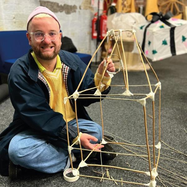 The image shows a person making a structure out of willow and tape. The structure in the shape of a house and they are holding it up and smiling at the camera.