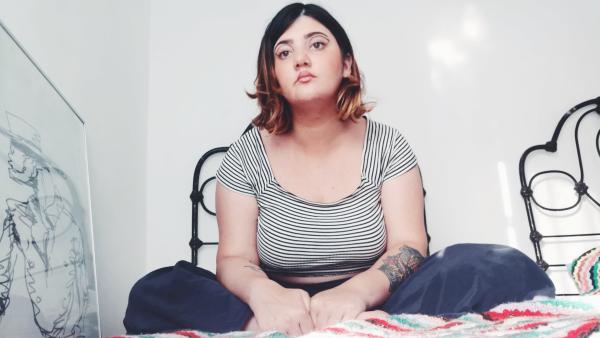 'Eeade', a young girl with brown hair and stylish eye makeup, sits on a bed cross-legged, looking into the distance