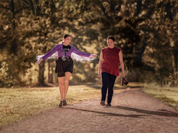 Two women walking down a path in a park, one wears a fringed cardigan and has her arms outstretched.