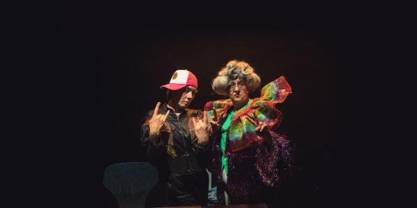 Two people are posing, one is wearing a white and red cap and the other a colourful dress. The background is black. 