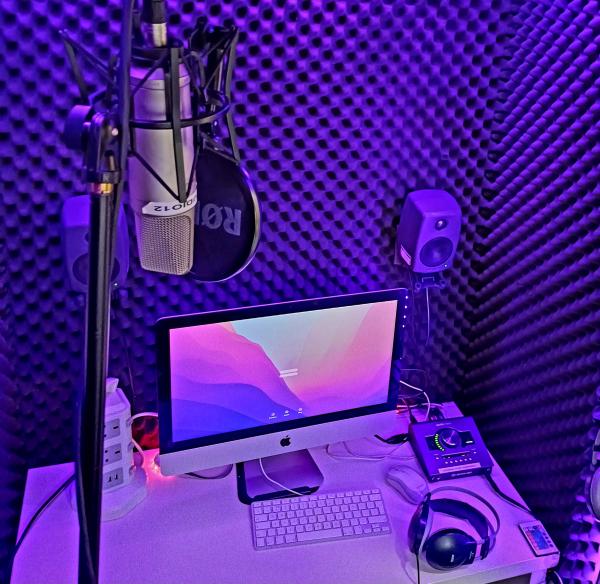 A computer, microphone, and headphones sit on a desk in a recording booth. Illuminated in purple light.