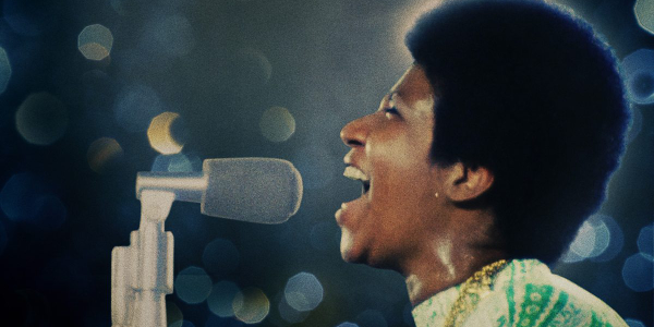 Aretha Franklin sings a powerful note into a microphone, soft out of focus lights in the background. 