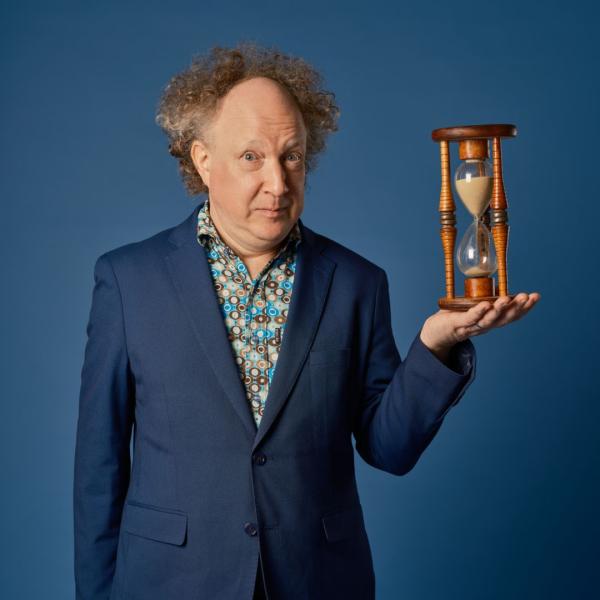 Andy Zaltzman in a sailor-blue suit jacket with a floral shirt under it holding a sand timer out in his left palm.
