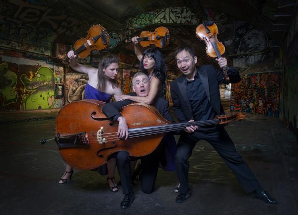 A group of four musicians in a tunnel covered in graffiti. There is a man holding a double bass kneeling down holding the instrument, to his right there is another man holding up his violin with a silly expression. There are two women next to him holding up their instruments in a playfully menacing way.