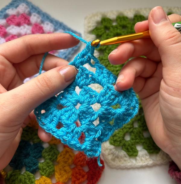 A blue crochet granny square wip, with two hands holding and crocheting it 