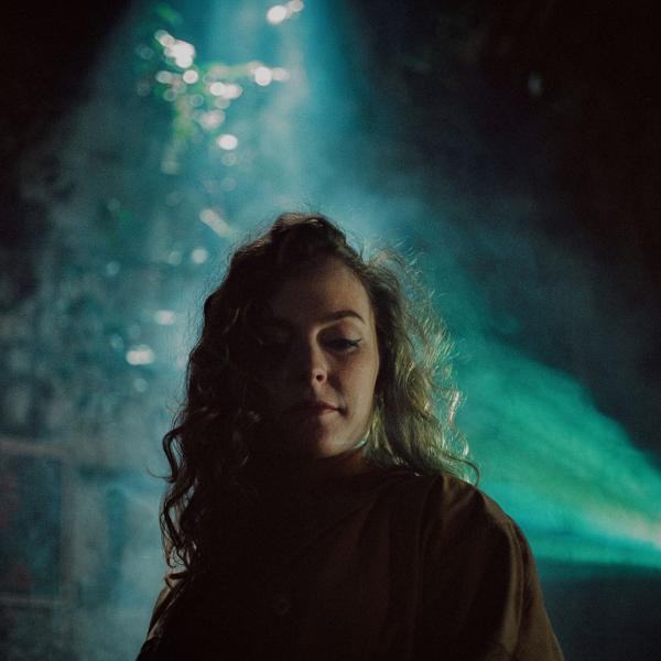 Shadowy image of Katherine Priddy looking down showing off the outline of her curly hair with a greeny-blue light lighting up a brick wall behind her.