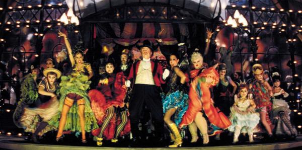 A stage full of colour can-can dancers with suited ringleader in the middle. 