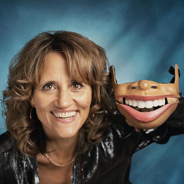 Image of Nina Conti's head against a blue background. She's holding up just a jaw with her left hand and she's mimicking it with her own mouth.