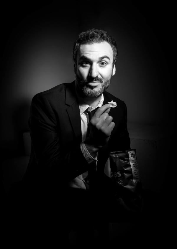 A picture of Patrick Monahan (the comedian) in black and white, holding a bag of 'cool original' flavoured Dorito crisps, looking into the camera inquisitively.