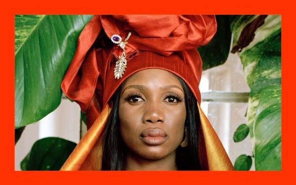 A woman, singer Rokia Kone, is wearing a green dress and orange head wrap and stood in front of leaves.