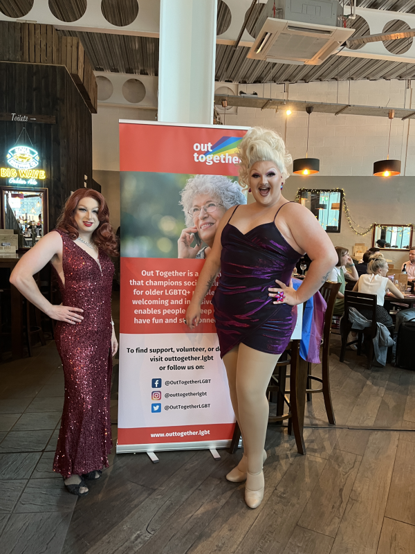 two drag queens posing next to a poster of the charity sponsor 